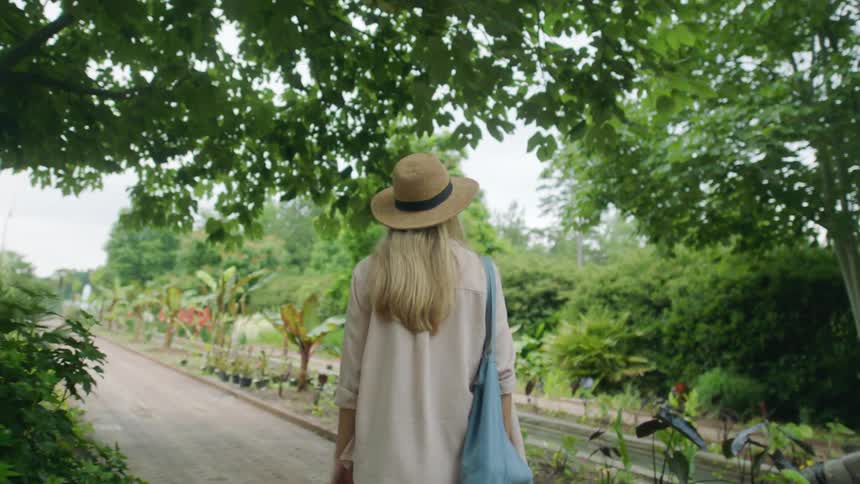 A woman with a hat and a bag walking on a pathway surrounded by trees and plants
