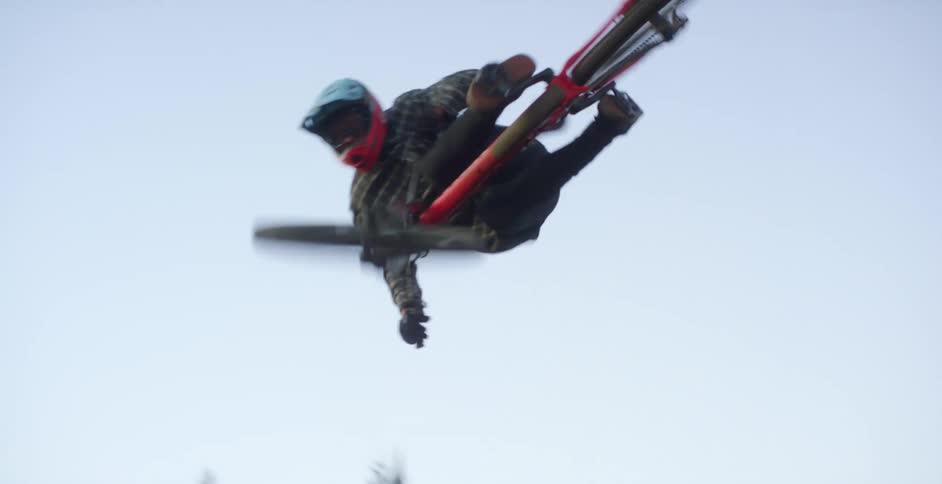 A cyclist goes airborne after riding up a dirt ramp in a clearing surrounded by forest during the day