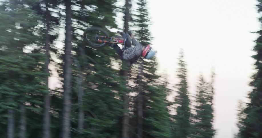 Man riding a red bike up a slope performs a flip in the air and lands it in a forest at daytime