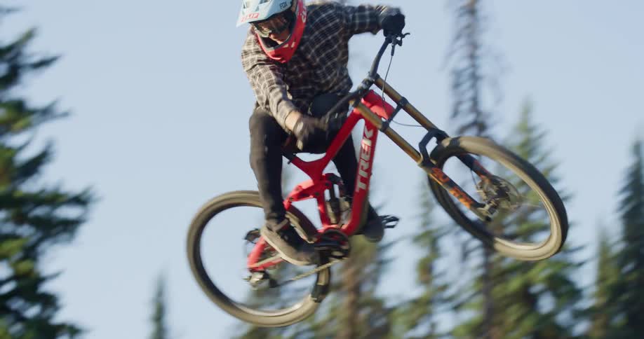 Cyclist rides a mountain bike jumping over a dirt ramp next to a pine tree forest