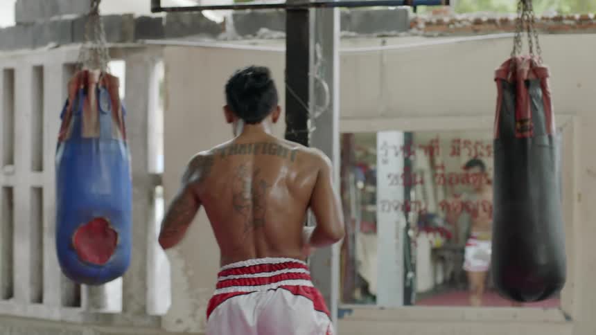 Young shirtless muscular man practices thai boxing in front of a mirror in a gym