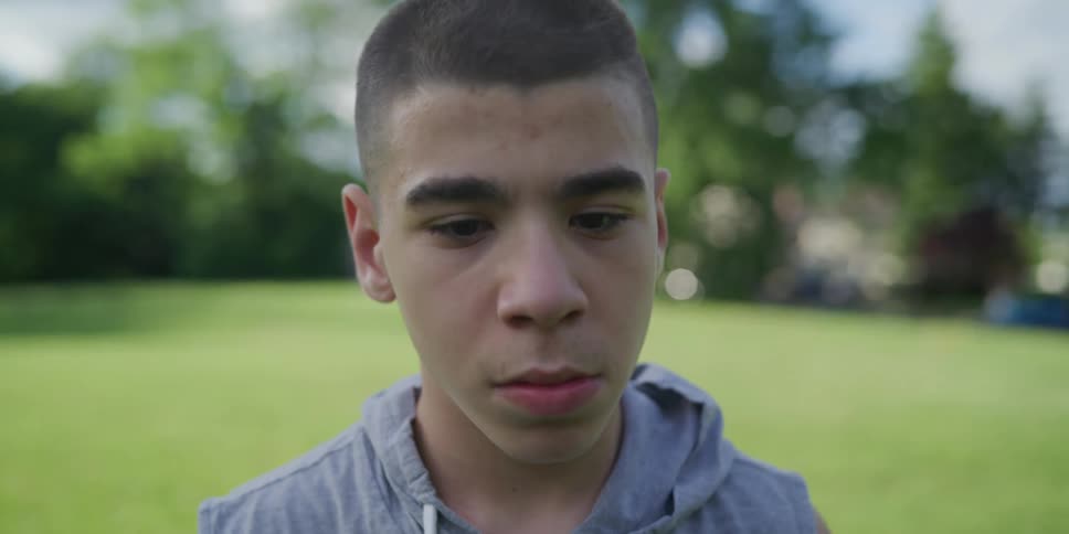 A young man standing in a field in a hoodie looks into the camera