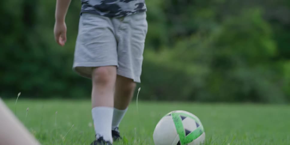 A young boy kicks a soccer ball while standing on the lawn outdoors during the day while another person lies on the grass