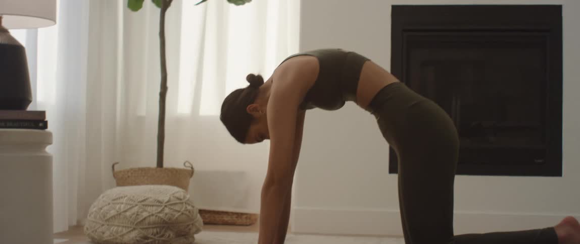 A young woman wearing close-fitting green yoga outfit alternates between cat and cow pose in a room with white walls and white curtains