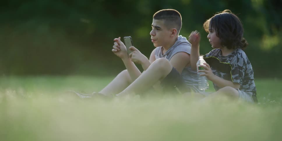 A male teenager texts on his phone as he sits with his brother on grass outside at dusk