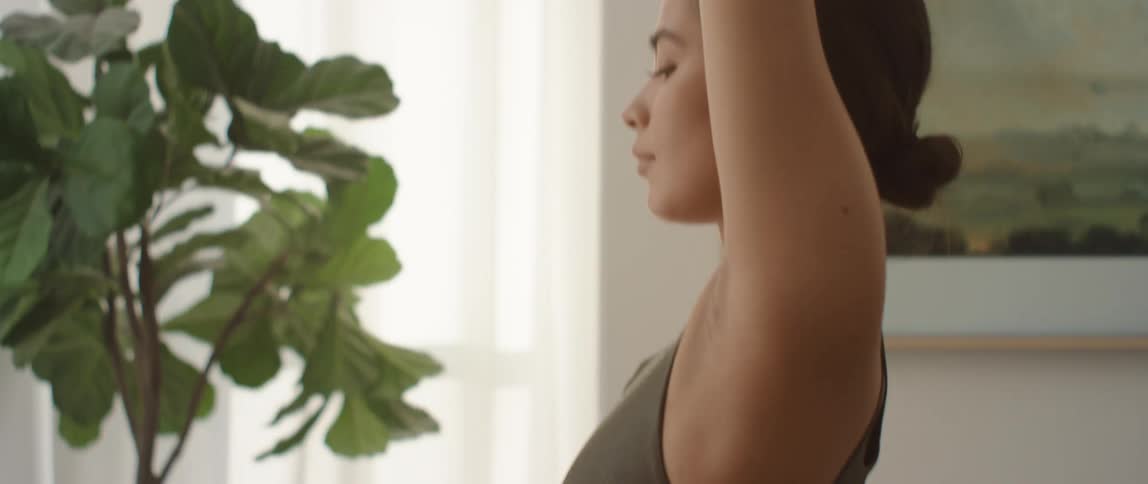 A woman standing in a room stretching with plants and windows in the background