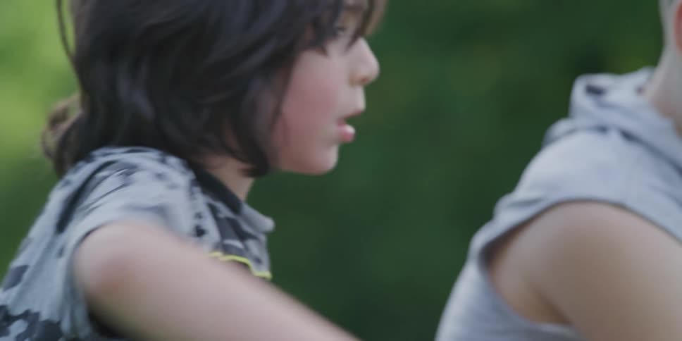 Two boys sit on a grassy field while looking at a smart phone as one of the boys kicks a soccer ball with trees in the background