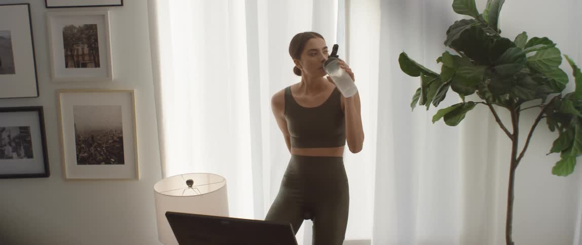 A young woman sitting on an exercise bike drinks from a water bottle while slowly pedaling