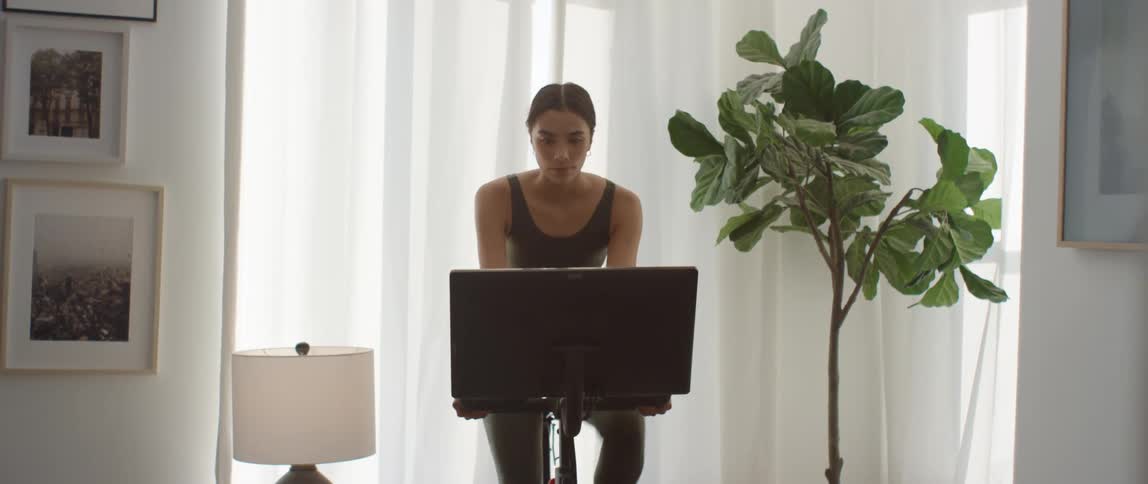A young woman pedals a stationary exercise bike while looking at its attached monitor in the living room of a house during the day