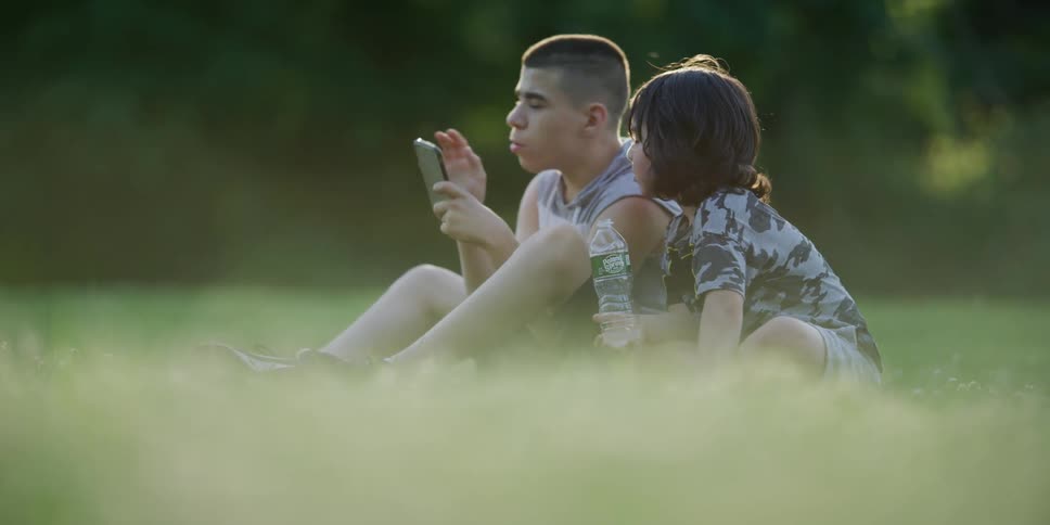 Two boys sitting on a grassy field looking at a smart phone with trees the sky and clouds in the background