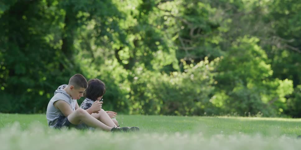 A boy kicks a soccer ball over to where his brother is sitting in the grass playing on a phone