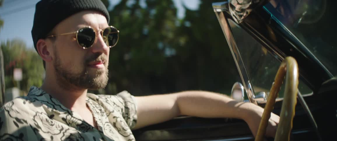 Man wearing knit hat and shades drives convertible with top down along streets