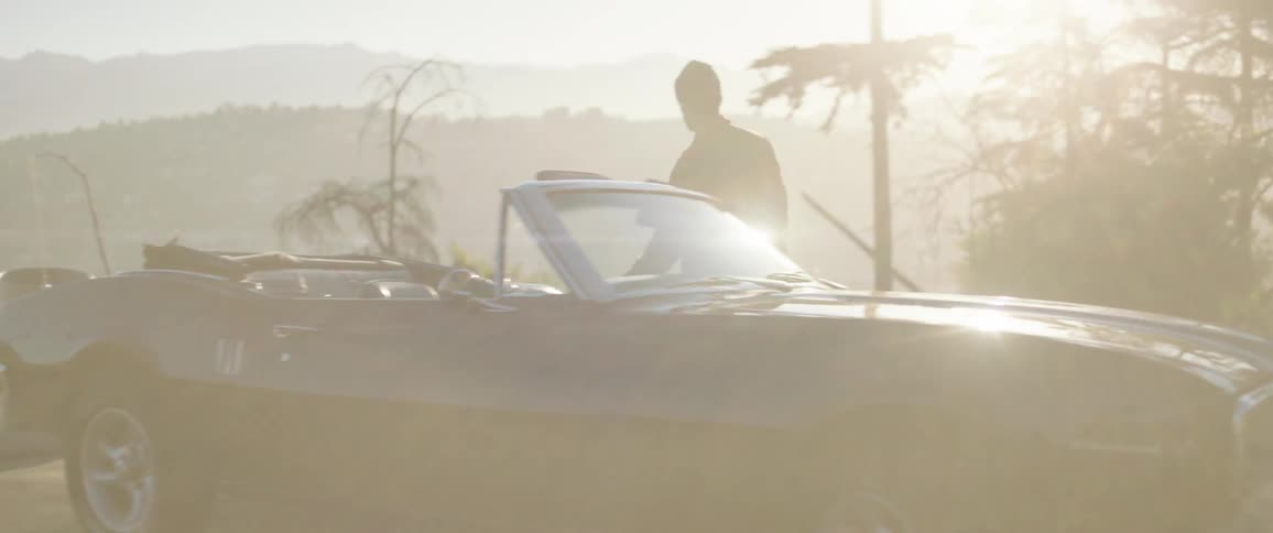 A man walks through a parking lot and enters his car outside with bright sunlight and mountains in the distance