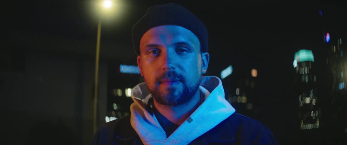 A man wearing a knot hat and a hoodie stands outdoors at night in a city area