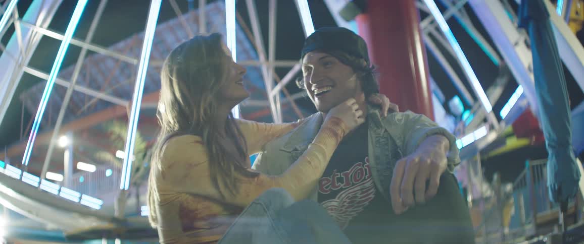 A white girl with long blonde hair is smiling with her arms around a white boy wearing a backwards baseball hat sitting together at a fairground