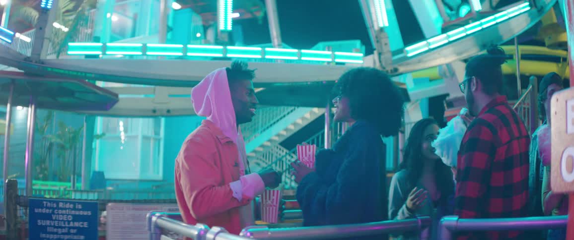 A man and a woman in an amusement park with the man feeding the woman popcorn with a ferris wheel in the background