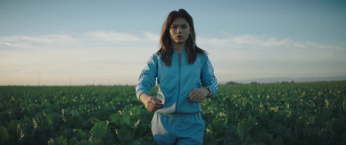 A young woman with dark brown hair is standing in a field of crops wearing a blue addidas tracksuit and a smart watch staring into the camera