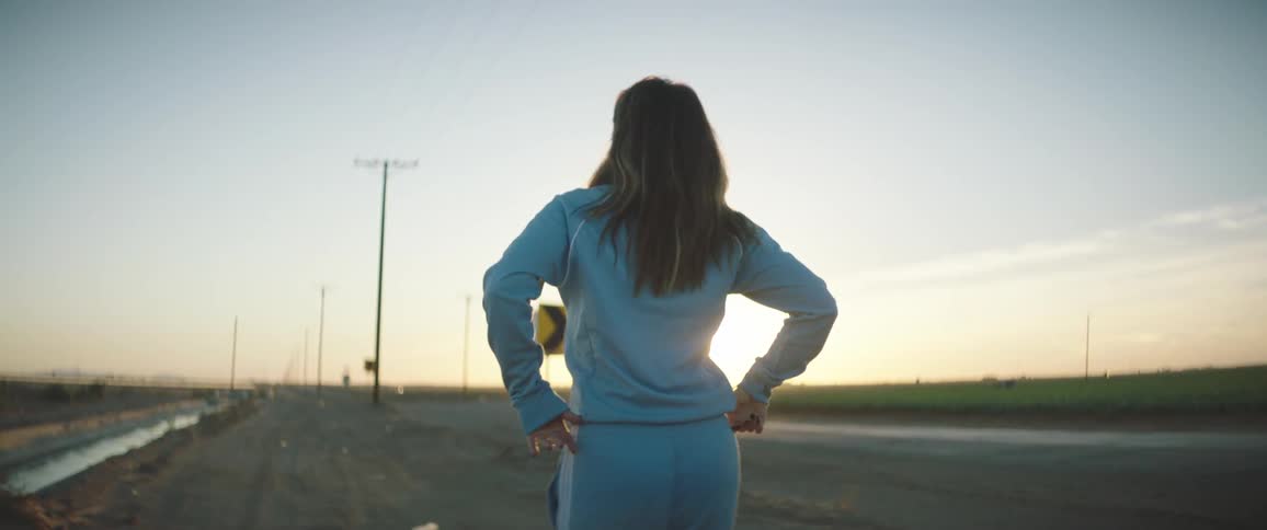 Woman walking down a road outdoors in front of a sunset looking back to pose at the camera