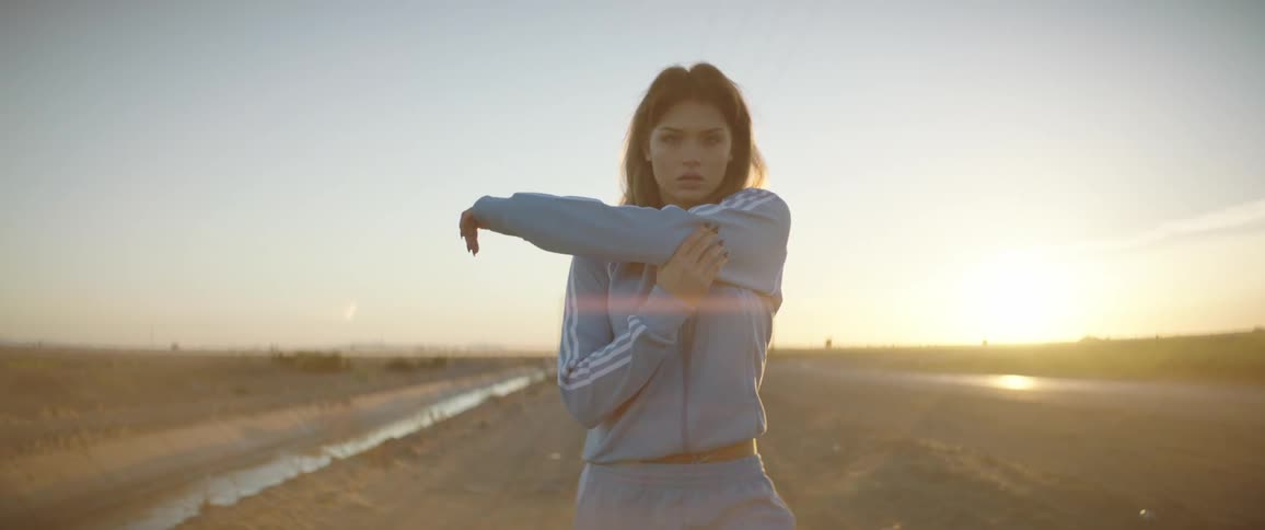 Young woman wearing sports clothes walking backwards on a field looking at the camera at sunset