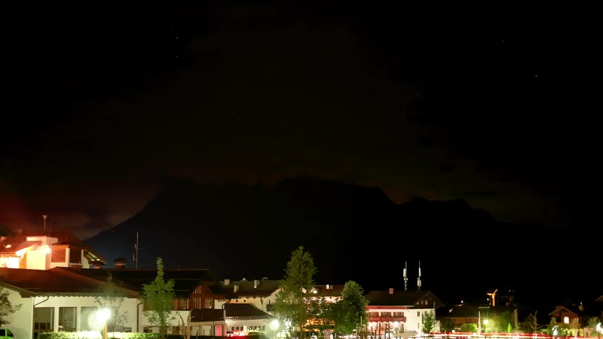 A urban neighborhood near a mountain with homes lit up under the night sky has cars coming and going through the night
