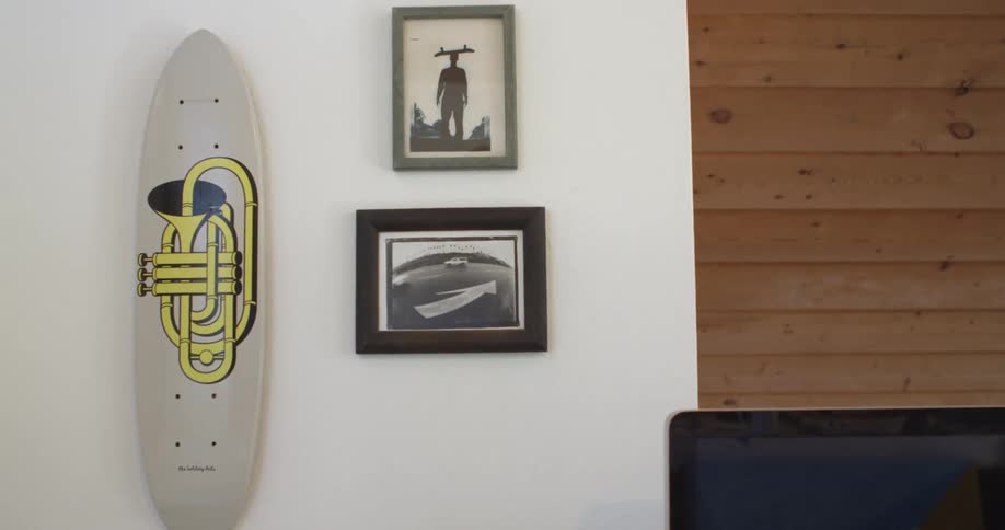 An interior wall of a bedroom has a mounted surfboard and two framed pictures displayed
