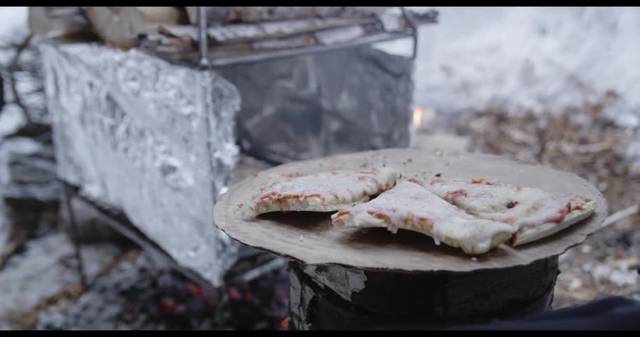 A person heats frozen pizza on a makeshift oven over a campfire at winter