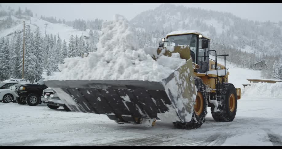 A loader carrying a huge scoop of snow drives through a snow filled parking lot