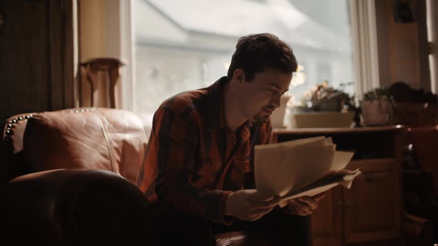 Man sitting on a small brown leather loveseat reading through pages of letters