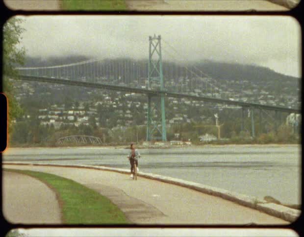 A woman bicycles along a path next to a river that passes beneath a suspension bridge
