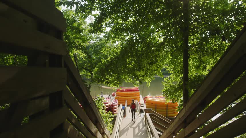 Young fit woman in sports clothing walking on a boardwalk towards a pier wint piles of kayaks on a river