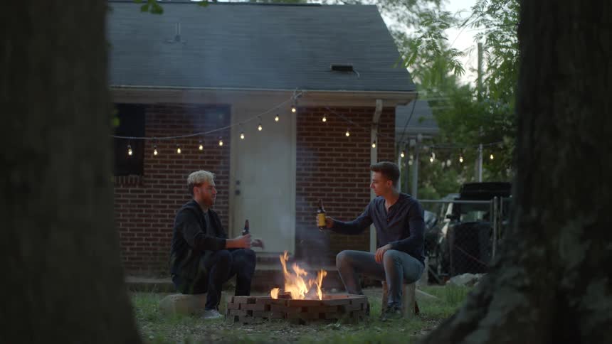 Two young men sit down at an outdoor fire pit and share bottles of beer