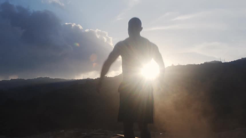 A shirtless man does jumping jacks in a dirt field near hills