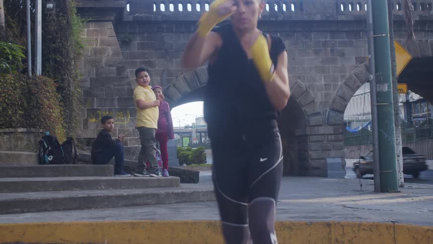 A woman runs on a city sidewalk punching the air like a boxer past small children sitting on the side of the road