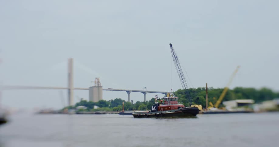 Boat drives in a river next to a shore with a long bridge and construction crane in the distance