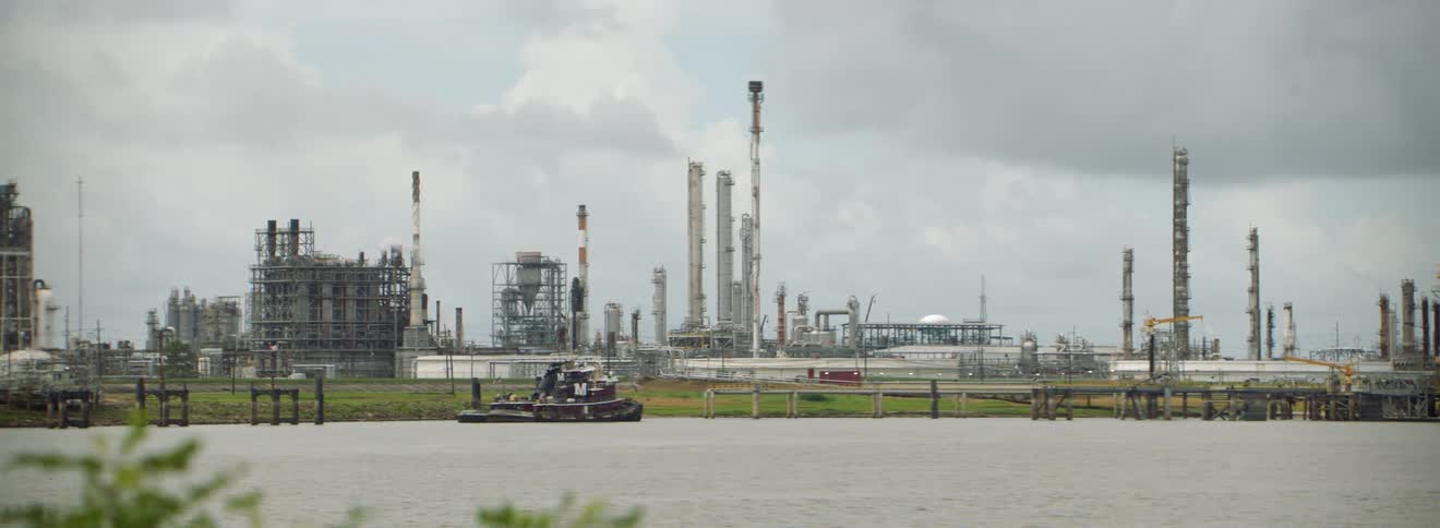 A large refinery facility is situated directly next to a river with a tugboat on it