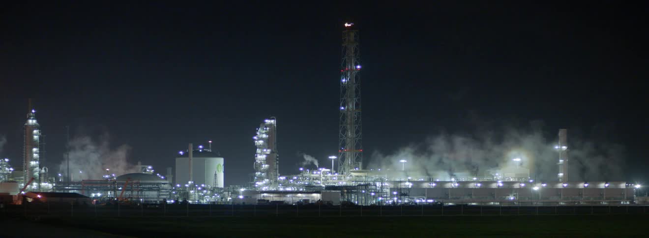 A large quantity of steam rises from a brightly illuminated refinery facility