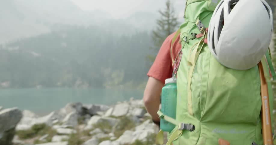 A man uses a walking stick to hike through boulders that lead to a forest and mountain along a lake