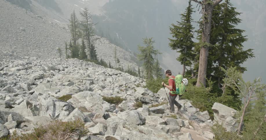A white man in a red shirt gray pants and green backpack is hiking with a walking stick in hand down a rocky steep mountainside