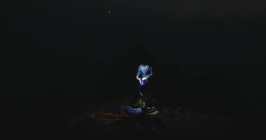 A man wearing a headlamp leans over and picks up items from his campsite