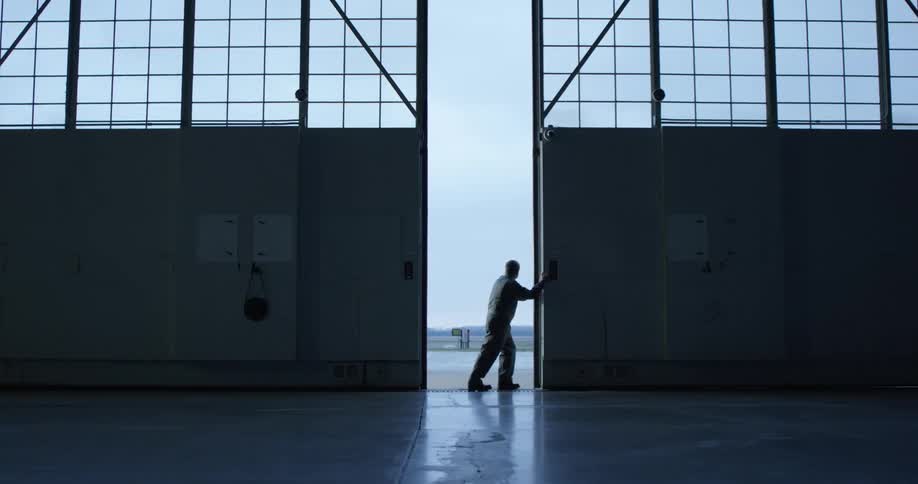 A man walks over to large hanger doors to slide it open to reveal an airfield