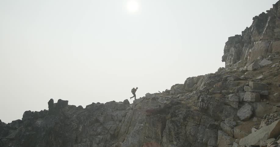 A hiker walks along an incline on a rocky cliff under a bright daytime sky