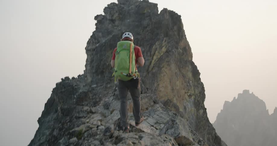 Man carrying a green backpack climbing to the peak of a mountain range