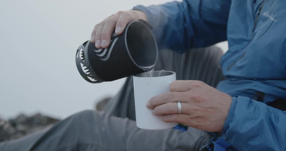 Man on a hike sitting over rocks pouring steaming hot water on a cup