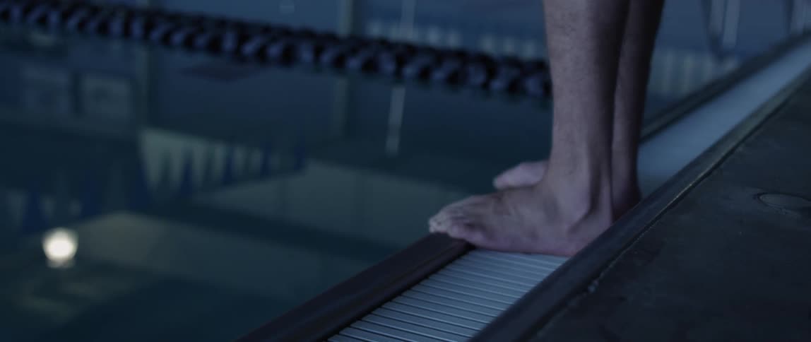 Ceiling light and banner are reflected on the surface of a swimming pool where a man stands on the edge