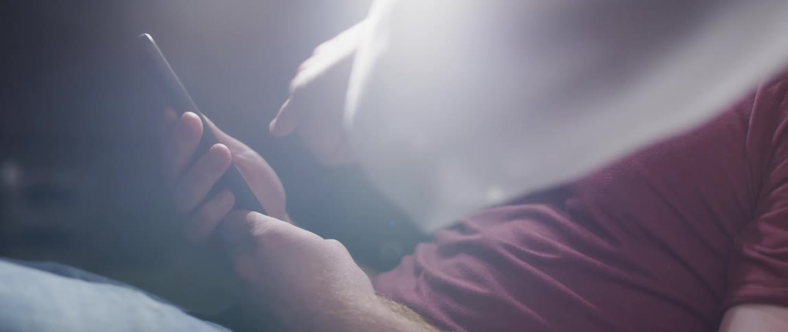 A white light beams down on a man on a smartphone while a hand with a long white sleeve reaches towards his screen