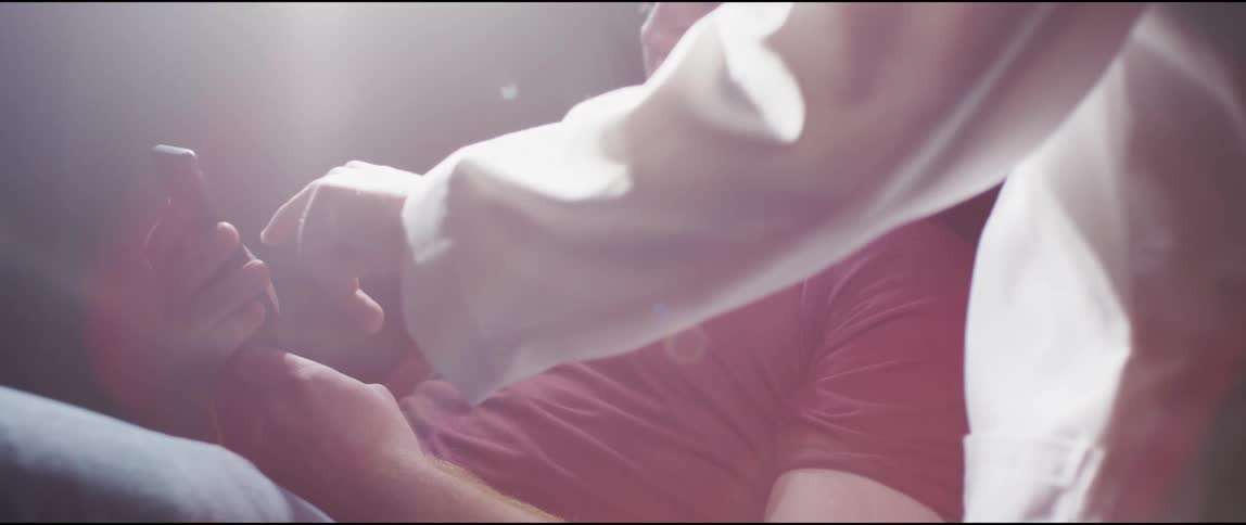 A man lies on a bed holding a cell phone as another person stands next to him and uses his hand to swipe on the screen