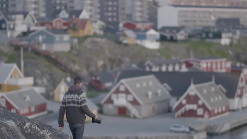 A man holding a camera walks down a hill towards a town