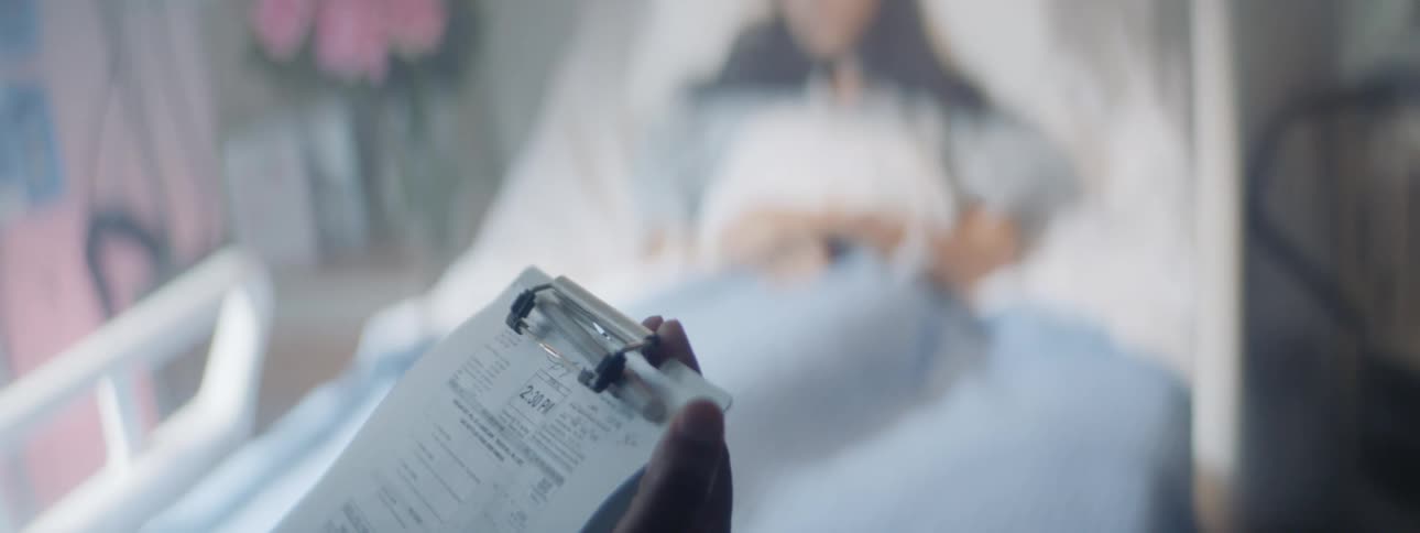 A person standing at the foot of a hospital bed with a clipboard as a woman lays on the bed