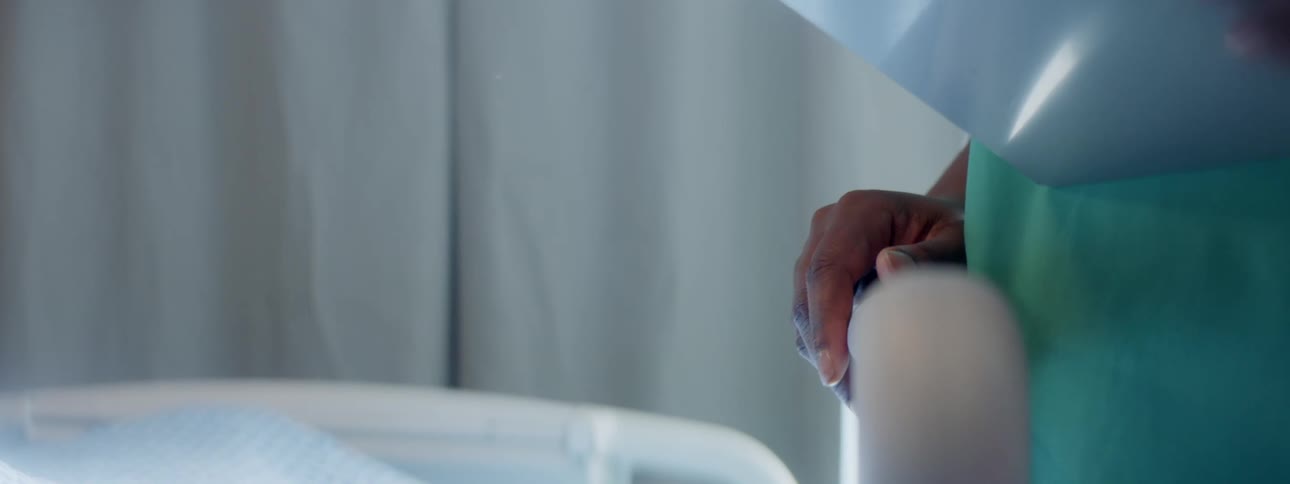 A nurse in teal scrubs holding a clipboard puts her hand on a hospital bed frame