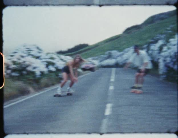 Man and a woman riding skateboards down a street on a hilltop next to mountain slopes at daytime